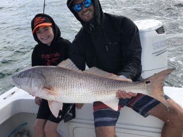A young angler and an adult proudly display a large Redfish while fishing on a boat, showcasing a successful day on the water.