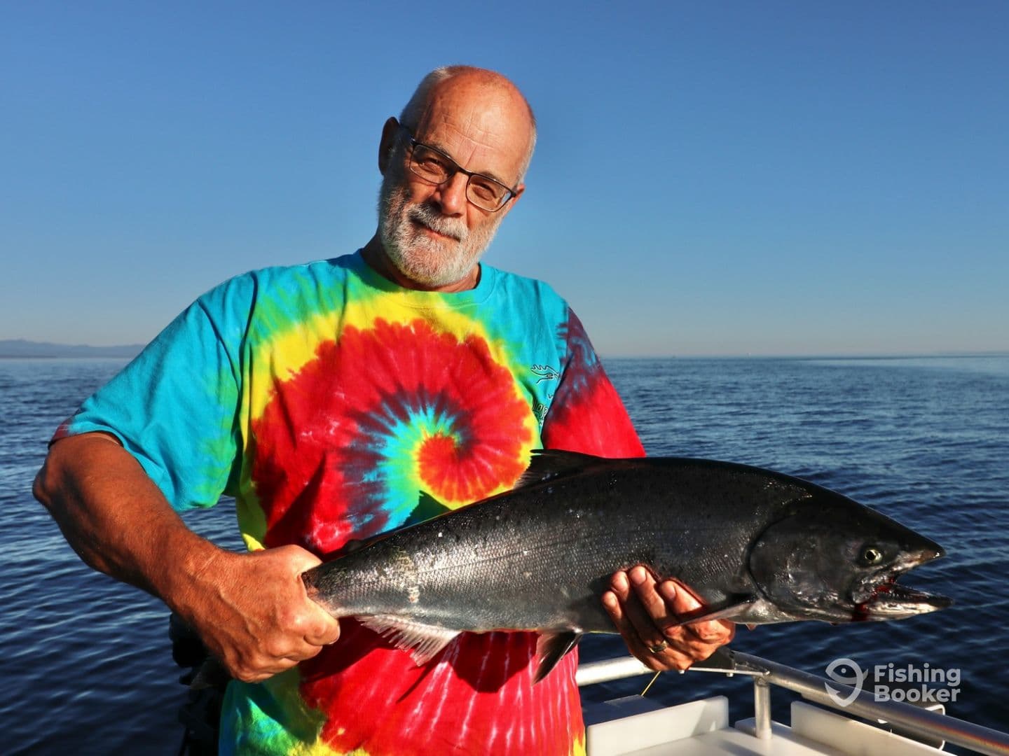 An angler proudly displaying a large Salmon while fishing in calm waters, showcasing a vibrant tie-dye shirt against a clear blue sky.