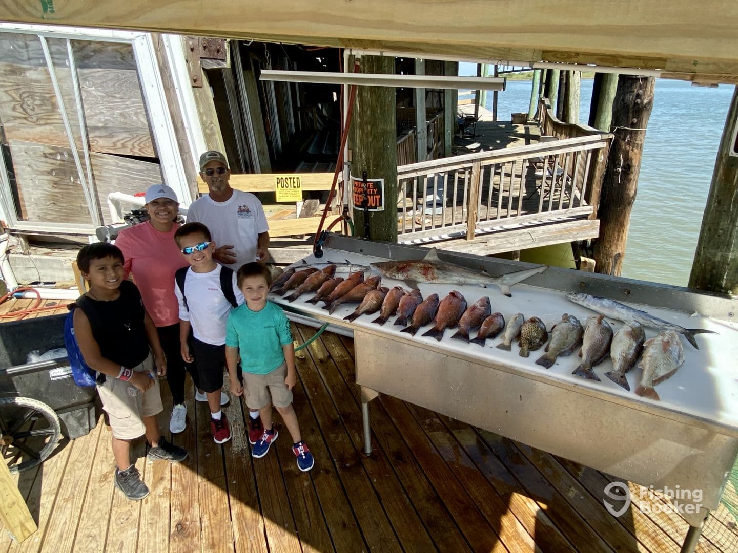 A family poses with their catch of the day, featuring a variety of fish displayed on a cleaning table at a dock.