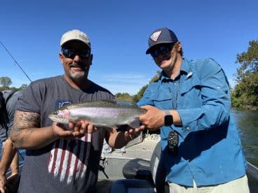 Two anglers proudly displaying a large Rainbow Trout while fishing on a scenic river.
