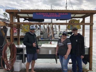 A group of anglers proudly posing in front of a catch board displaying several fish, including Speckled Trout, at a fishing dock.