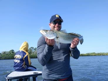 An angler proudly displays a large Speckled Trout while fishing on a boat, with a young companion in the background.