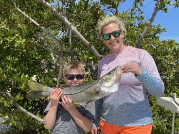 A young boy and his mother proudly display a large Snook caught during a family fishing trip in a lush coastal environment.