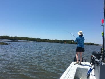 An angler casting a line from a small boat in a serene coastal environment, surrounded by lush greenery.