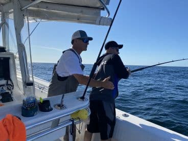 Two anglers engaged in fishing on a sunny day, with one assisting the other as they reel in a catch from the deep blue waters.