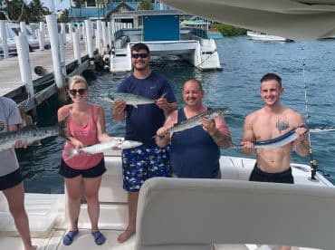 Group of anglers proudly displaying their catch, including a Barracuda and a Mackerel, aboard a fishing boat in a tropical marina setting.