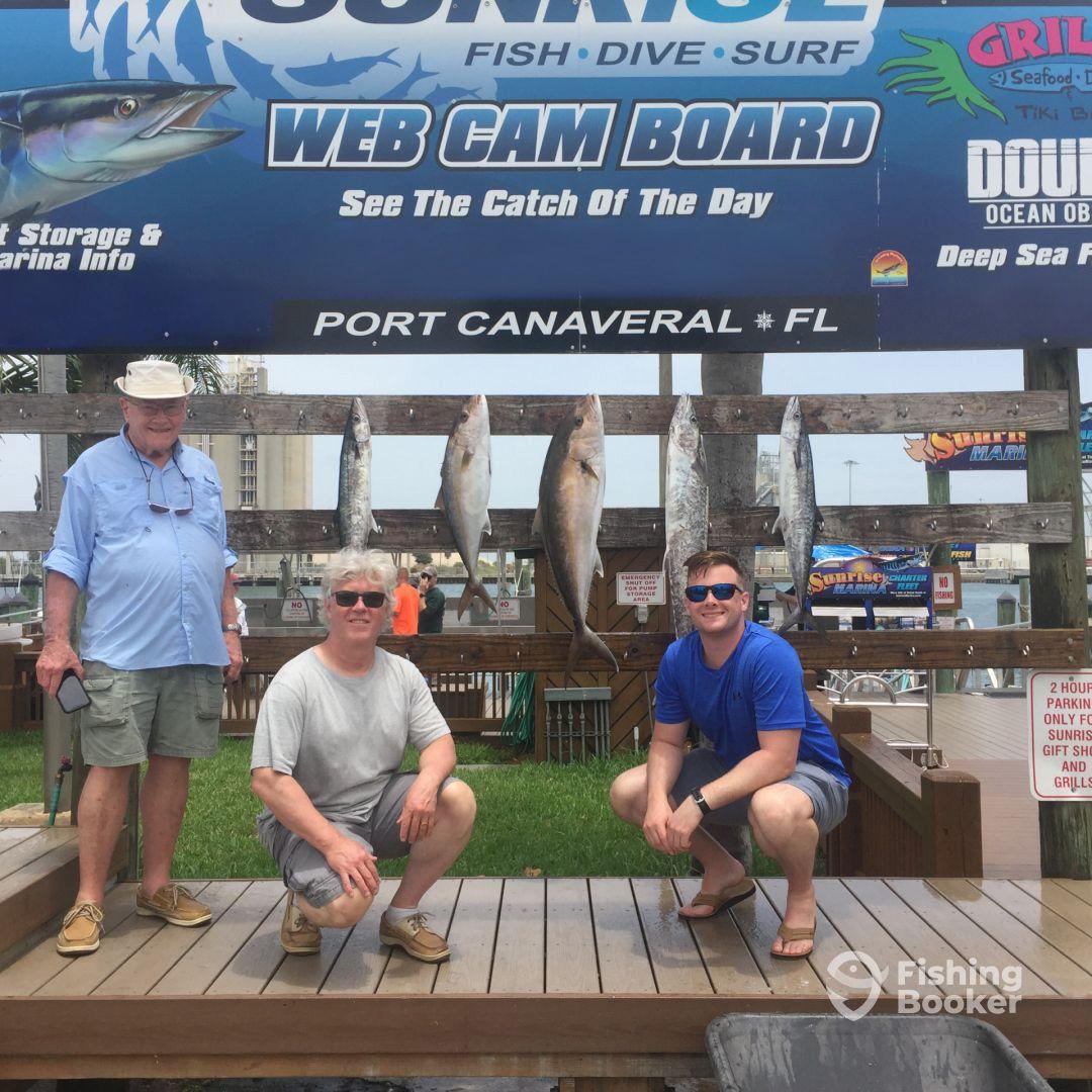 Three anglers posing in front of a catch board displaying several fish, including King Mackerel, at Port Canaveral, showcasing a successful fishing trip.
