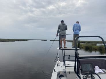 Two anglers fishing from a boat in a calm waterway, surrounded by lush marshland, showcasing a serene fishing environment.