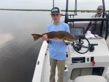 A young angler proudly displaying a Redfish while aboard a fishing boat in a calm waterway.