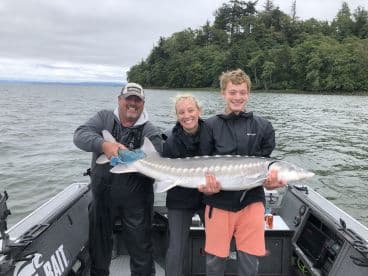 Family group proudly displaying a large Sturgeon caught during a fishing trip, with a scenic backdrop of lush greenery along the water.