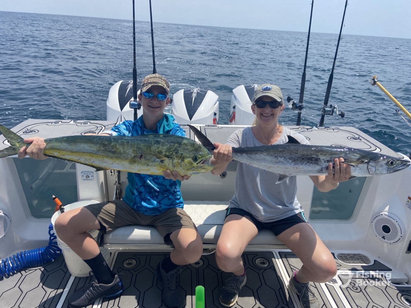 A young angler and an adult proudly displaying their catches, a Mahi Mahi and a Kingfish, aboard a fishing boat in open waters.