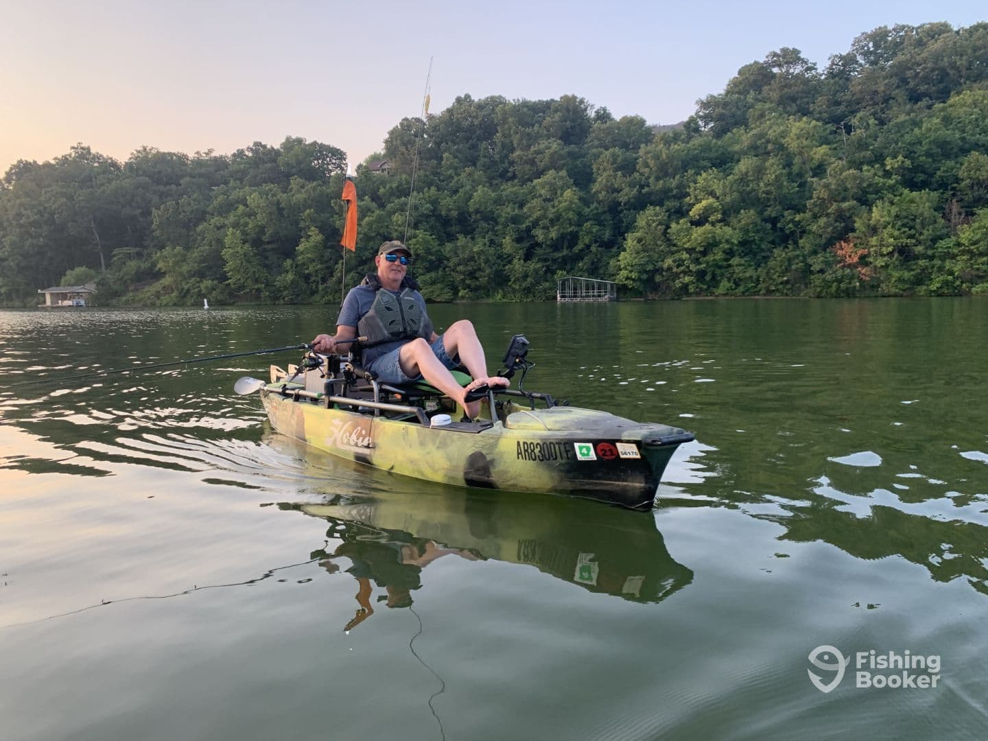 A solo angler enjoying a peaceful morning on a Hobie kayak, casting lines in a serene lake surrounded by lush greenery.