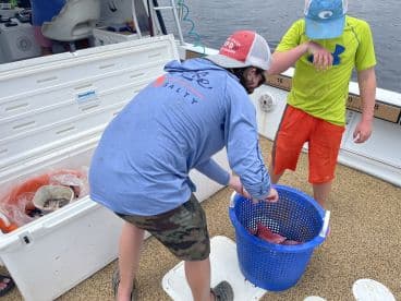 Two young anglers on a boat examining a blue basket filled with freshly caught fish, showcasing a fun day of fishing.