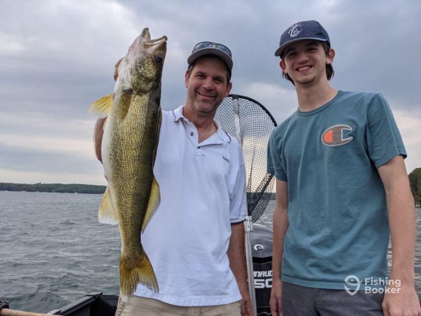 A proud angler displays a large Walleye while fishing with a young companion on a scenic lake.