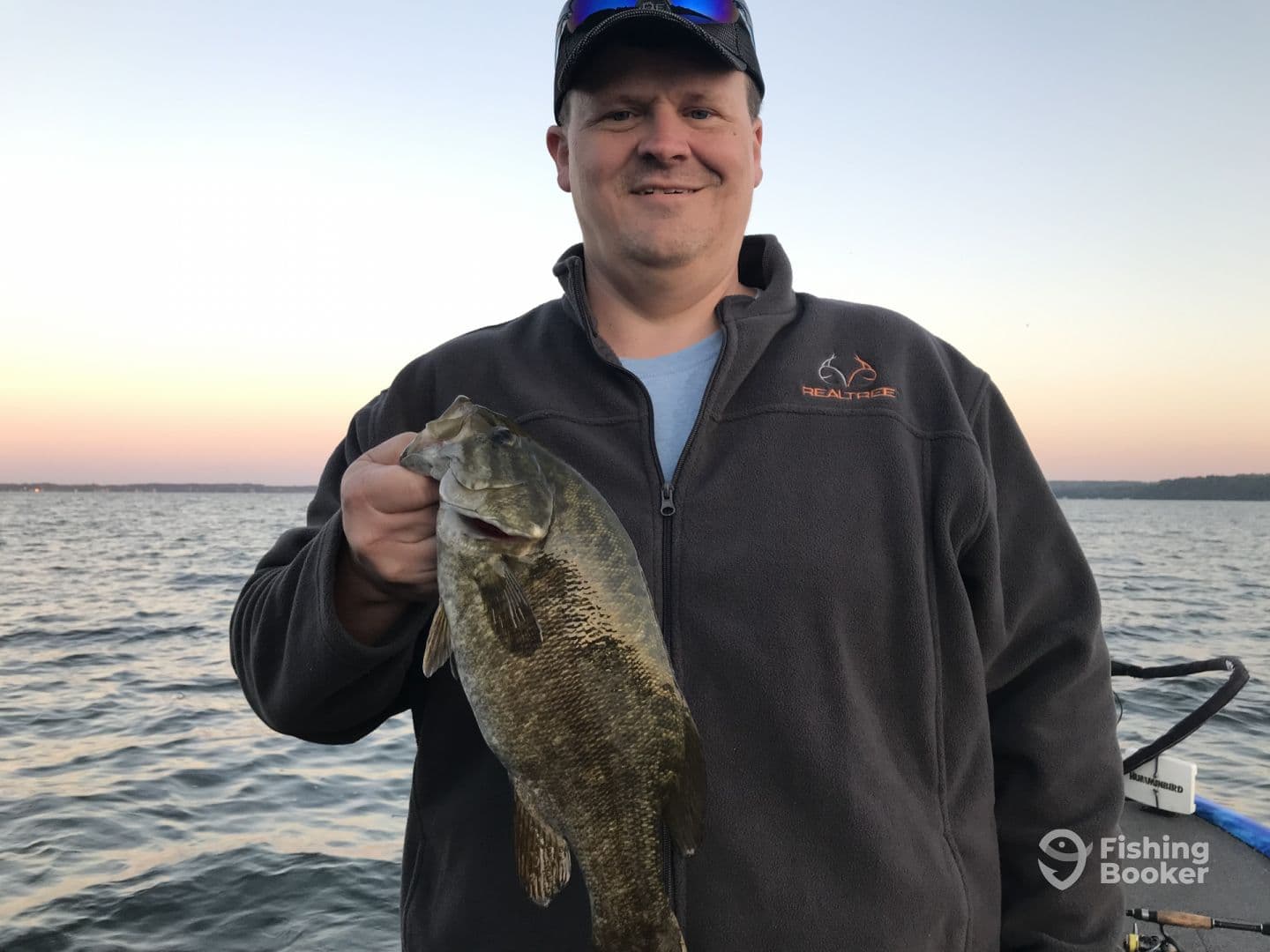 An angler proudly displaying a sizable Smallmouth Bass while fishing on a serene lake during sunset.