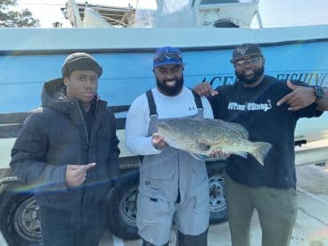 Three anglers proudly displaying a large Grouper after a successful fishing trip, with a fishing charter boat in the background.