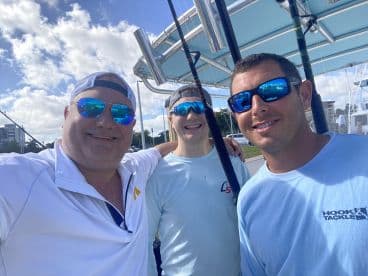Three anglers posing for a selfie on a fishing boat, showcasing a sunny day and fishing gear in the background.