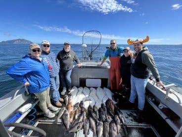 A group of anglers proudly posing on a fishing boat with a successful catch of various fish, including halibut and rockfish, against a scenic ocean backdrop.