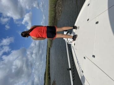 A young woman stands on the side of a boat, enjoying the scenic view of the marshland under a partly cloudy sky.