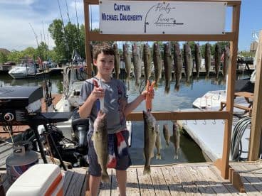 A young angler proudly displaying a large Walleye at the dock, surrounded by a successful catch of multiple fish on a sunny day.