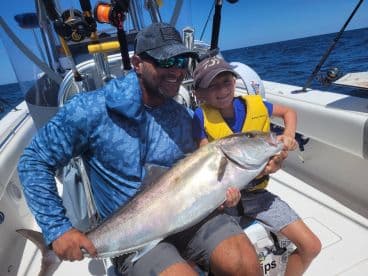 A young angler and an adult proudly displaying a large fish on a boat during a sunny offshore fishing trip.