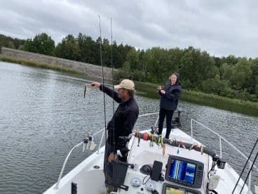 A young girl and an adult angler are fishing on a boat, with the adult proudly displaying a catch while the girl prepares to reel in her line in a serene freshwater setting.