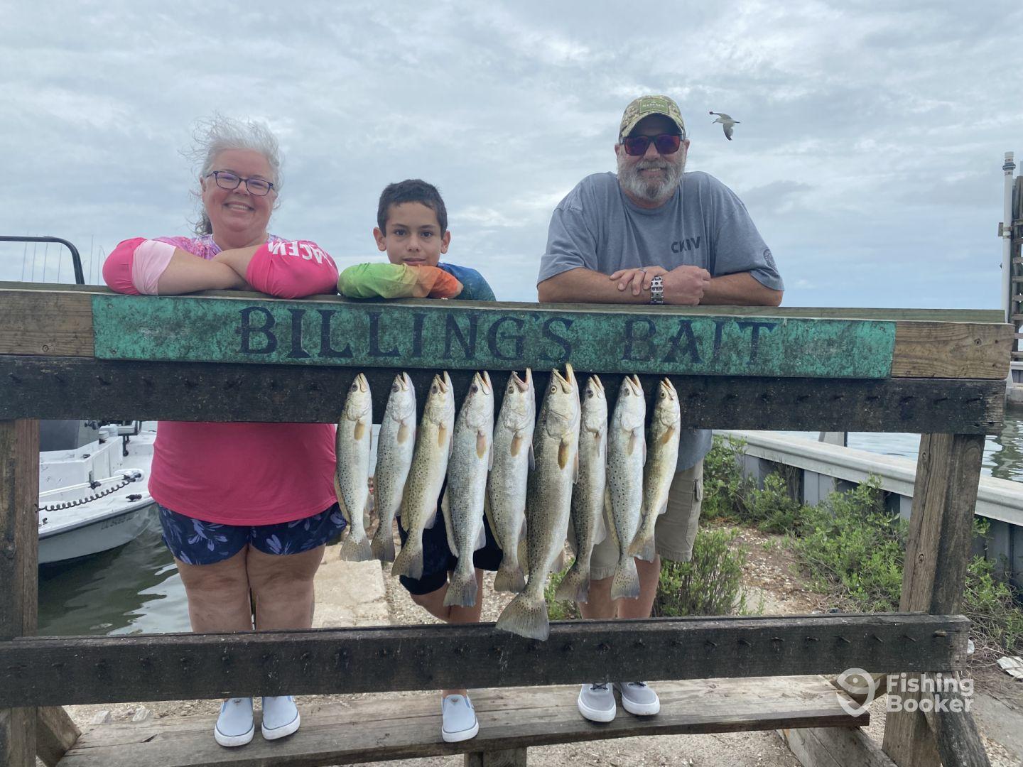A family proudly displays their catch of Speckled Trout on a catch board at a fishing dock, showcasing a successful day on the water.