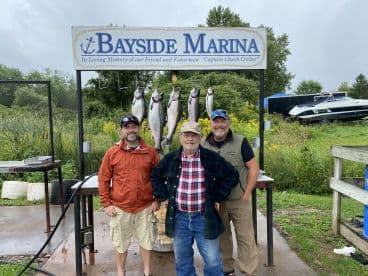 Three anglers proudly posing in front of a catch board displaying several Salmon at Bayside Marina, showcasing a successful fishing trip.