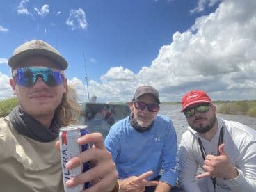 Three anglers enjoying a day on the water, seated inside a boat with a scenic backdrop of clouds and marshland.
