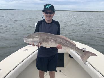 Young angler proudly holding a large Redfish while on a boat in a coastal fishing environment.