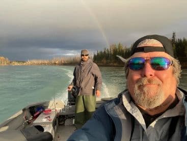 Two anglers enjoying a scenic boat ride on a river, with a rainbow in the background, showcasing a relaxed fishing environment.