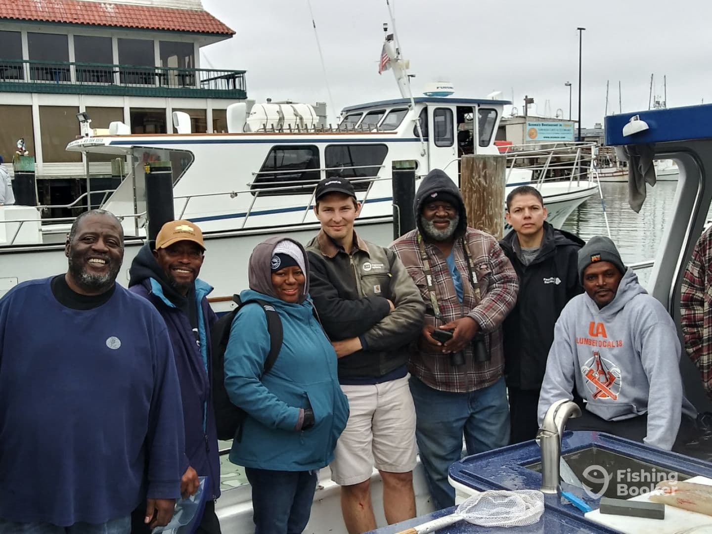 A diverse group of anglers posing together on the boat after a fishing trip, with multiple boats visible in the background at the marina.