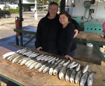 A couple proudly poses with a neatly arranged catch of Speckled Trout on a cleaning table after a successful fishing trip.