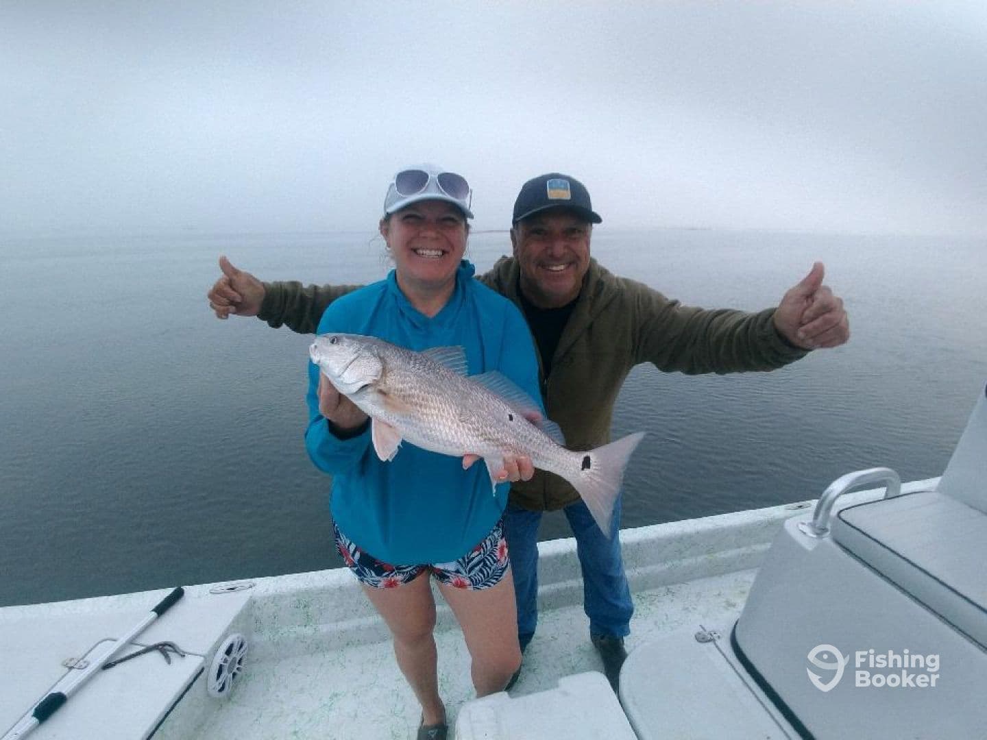 A happy angler proudly displaying a Redfish while fishing on a foggy day, with a fellow angler giving a thumbs up in the background.