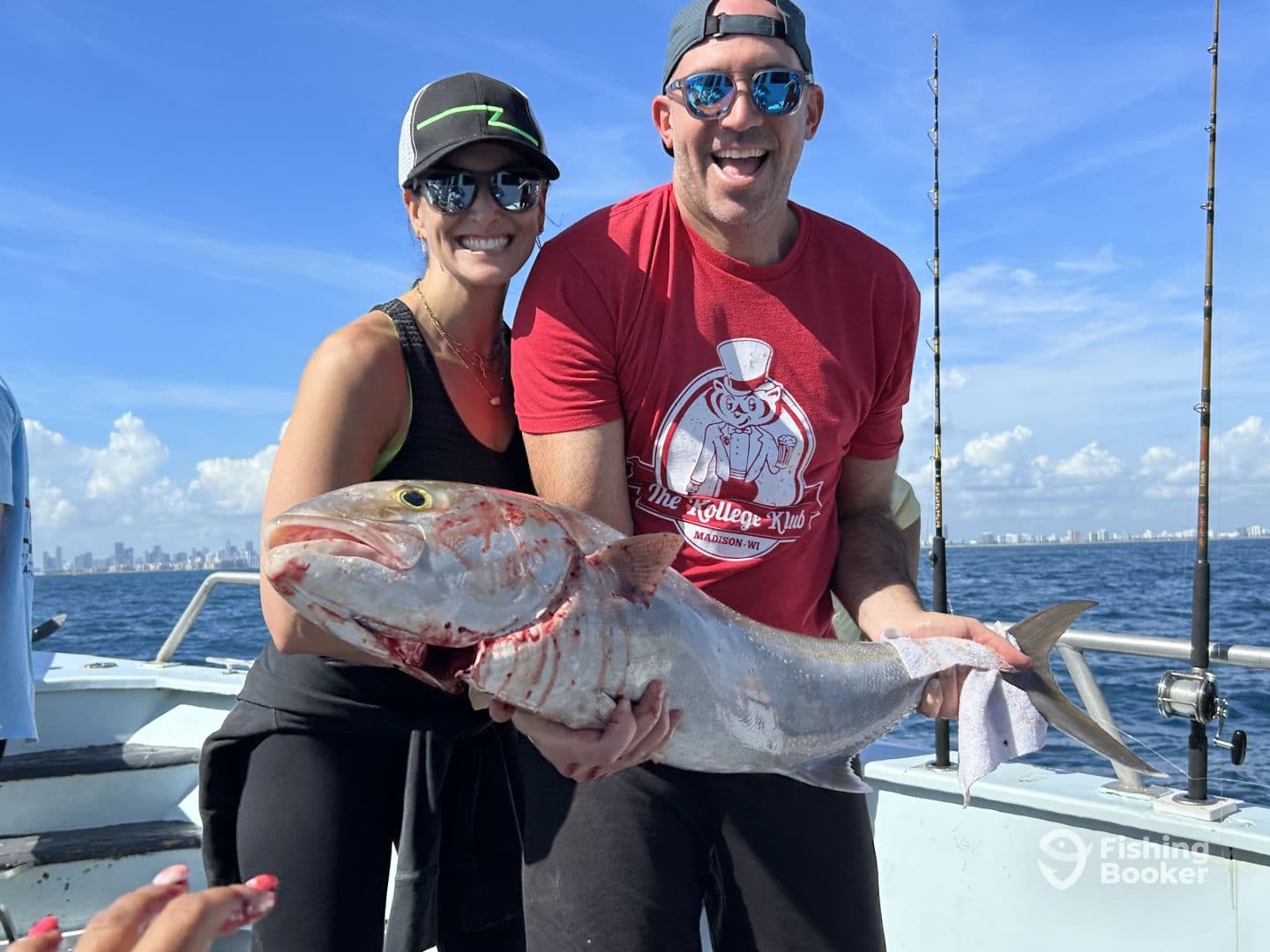 Two anglers proudly displaying a large Grouper while fishing off the coast, with a city skyline in the background.