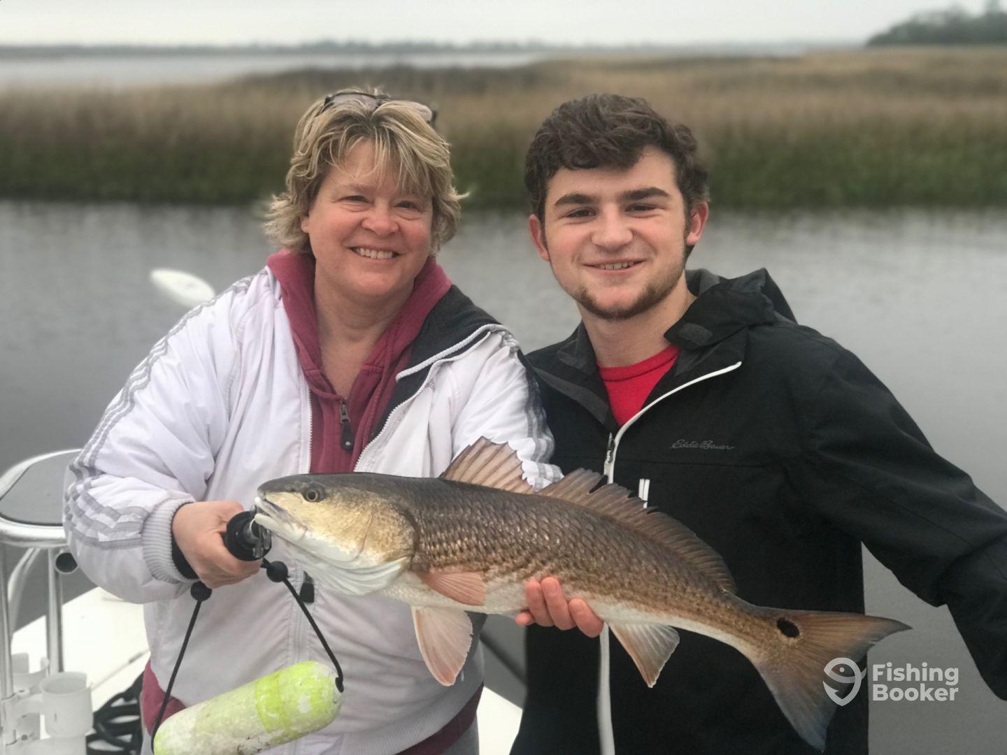 A mother and son proudly displaying a large Redfish while fishing in a scenic coastal environment.