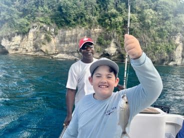 A young angler proudly displaying a small fish while fishing on a boat, with a scenic rocky backdrop.