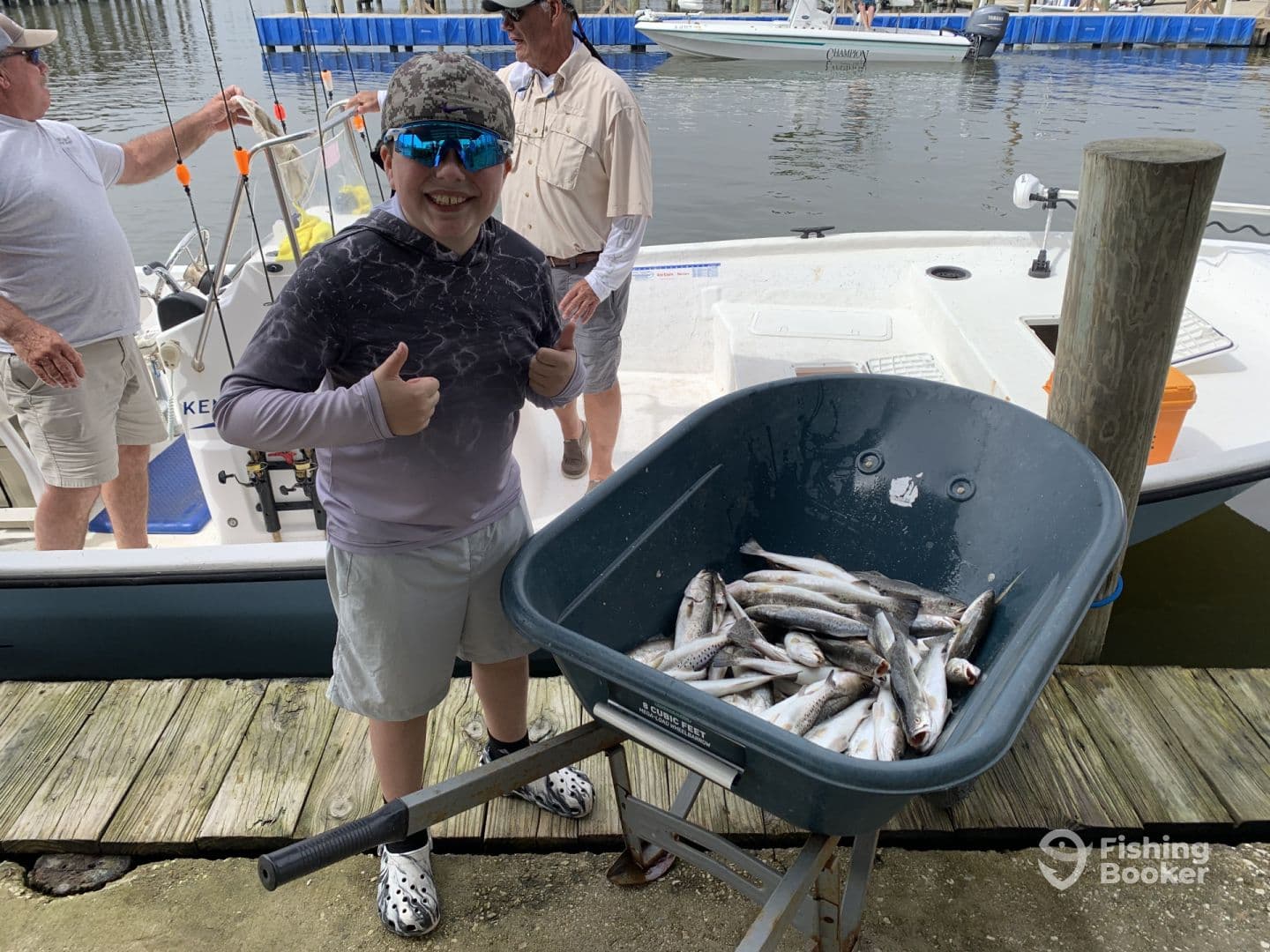 A young angler gives a thumbs up next to a wheelbarrow filled with freshly caught fish at the dock, showcasing a successful fishing trip.