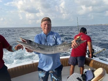 An angler proudly displays a large Barracuda while fishing on a charter boat, surrounded by a beautiful ocean backdrop.