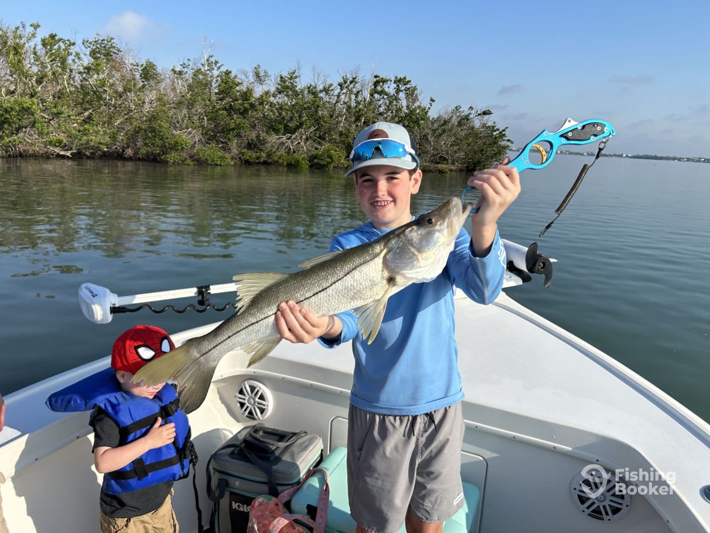 A young angler proudly displaying a Snook caught while fishing on a boat, with a scenic backdrop of mangroves.