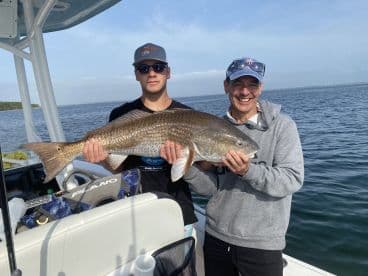 Two anglers proudly displaying a large Redfish while aboard a fishing boat, enjoying a successful day on the water.