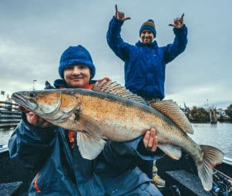 A young angler proudly displaying a large Walleye while fishing on a boat, with an adult celebrating in the background.