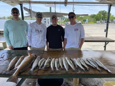 A group of four anglers proudly displaying their catch of Redfish and Speckled Trout on a catch board at the dock, showcasing a successful day of fishing.