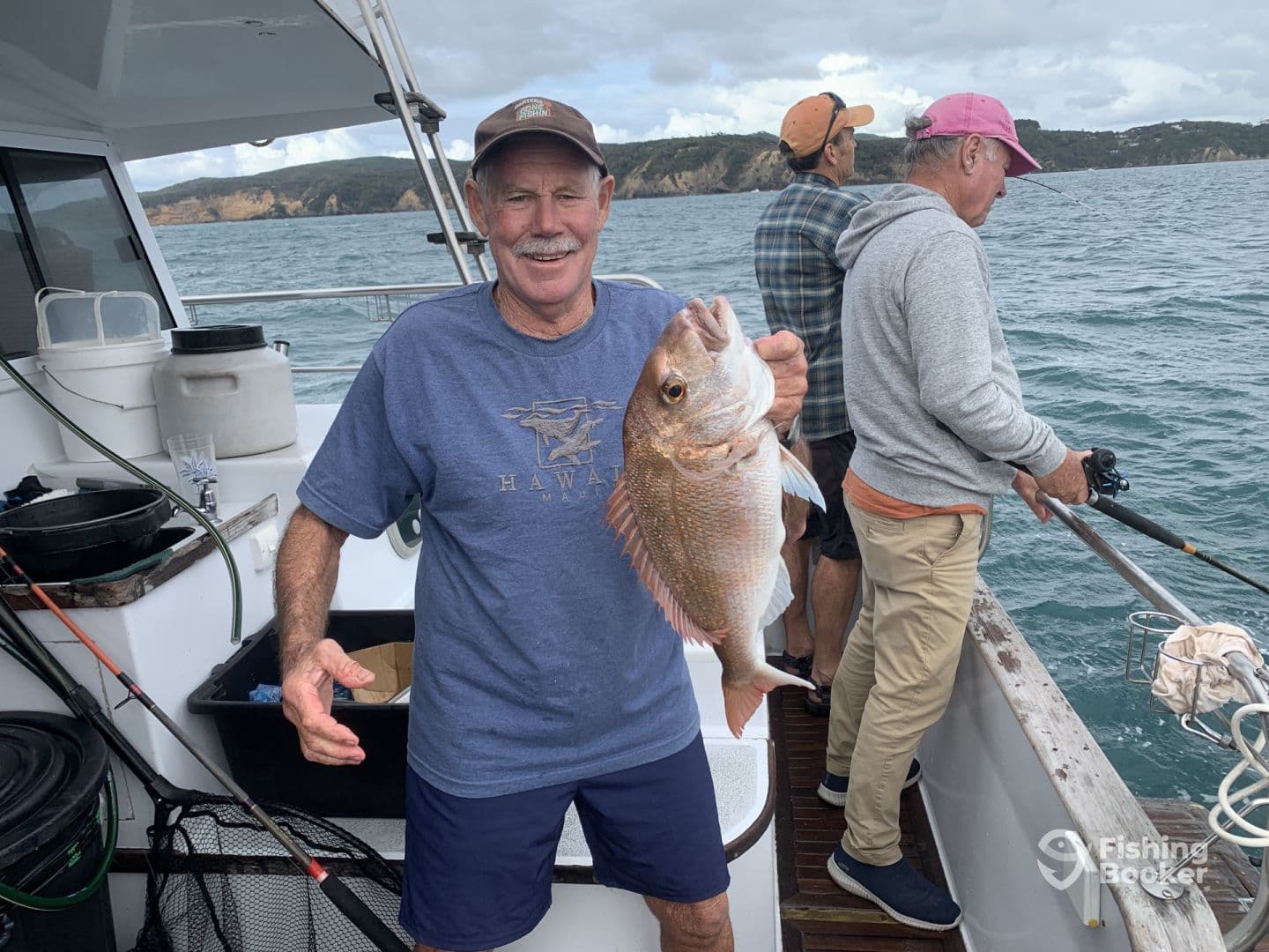 An angler proudly displaying a Snapper caught while fishing on a charter boat, with fellow fishermen in the background enjoying the day on the water.
