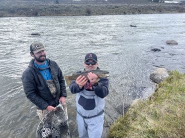 Two anglers proudly displaying a caught Brown Trout while wading in a river, showcasing a successful day of fly fishing.