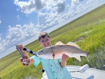 Young angler proudly holding a large Redfish while fishing in a grassy coastal area.