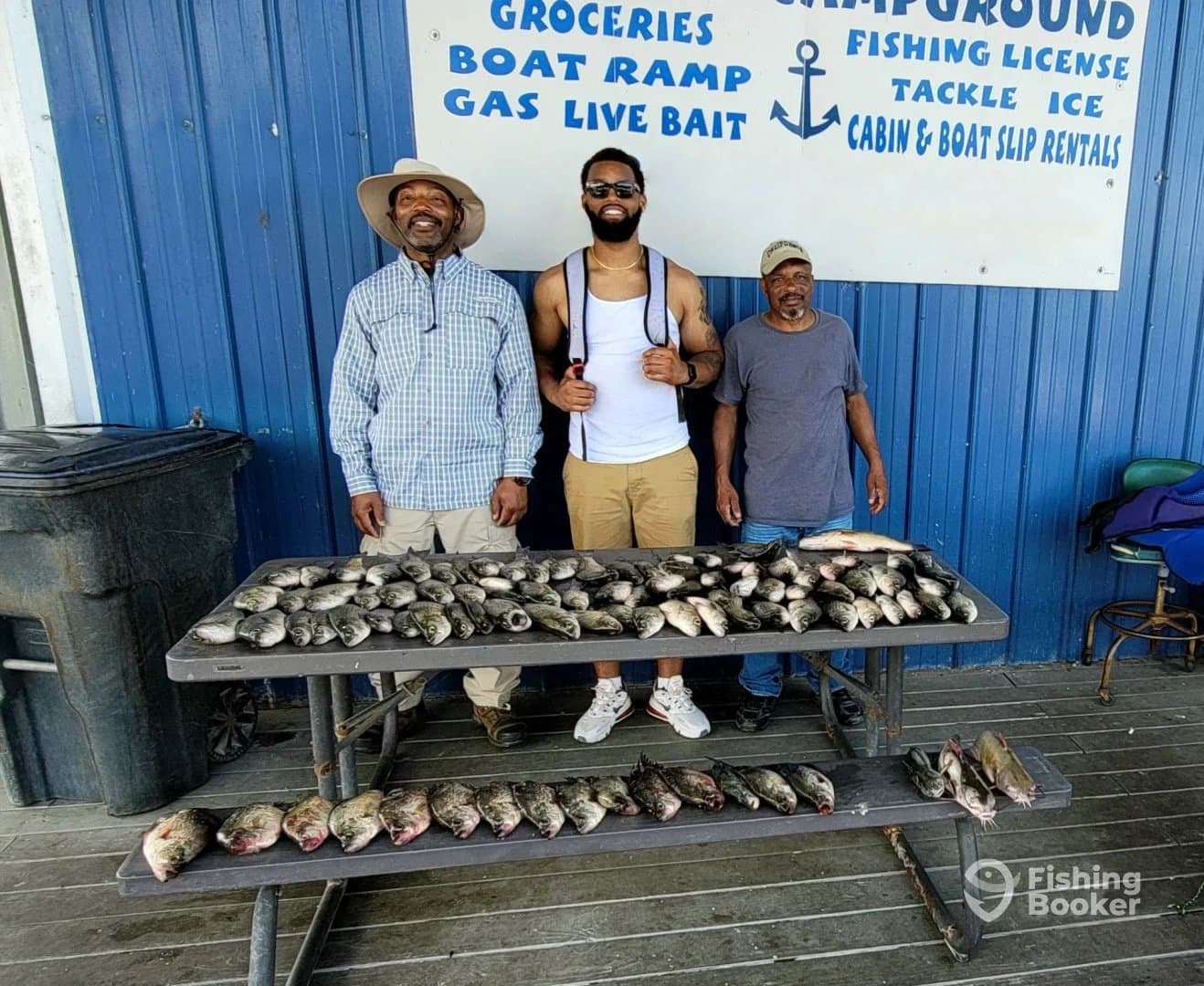 Three anglers proudly displaying their catch of various fish on a table at a fishing dock, showcasing a successful day on the water.