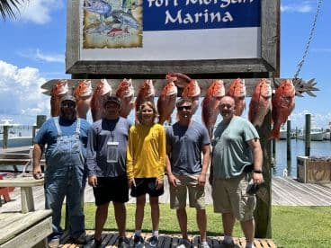 A group of anglers proudly posing in front of a catch board displaying several Red Snapper at Fort Morgan Marina, showcasing a successful fishing trip.