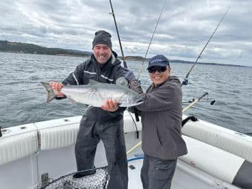 Two anglers proudly displaying a large Salmon while fishing on a boat in a scenic coastal environment.