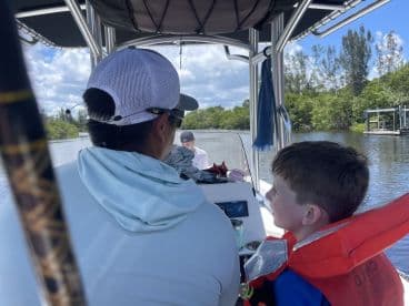 A young boy wearing a life jacket sits beside an adult on a boat, enjoying a scenic view of the waterway surrounded by lush greenery.